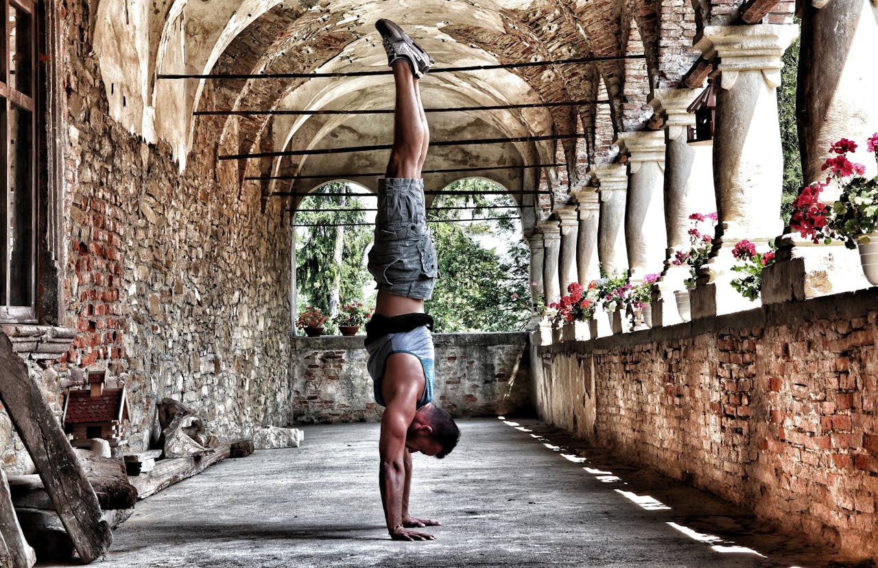 A man performs a handstand in a rustic, arched corridor with brick walls and flowers.